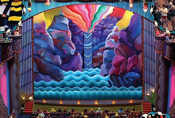 The image depicts a colorful stage design with a canyon backdrop, floral arches, spotlights and a performance setting.