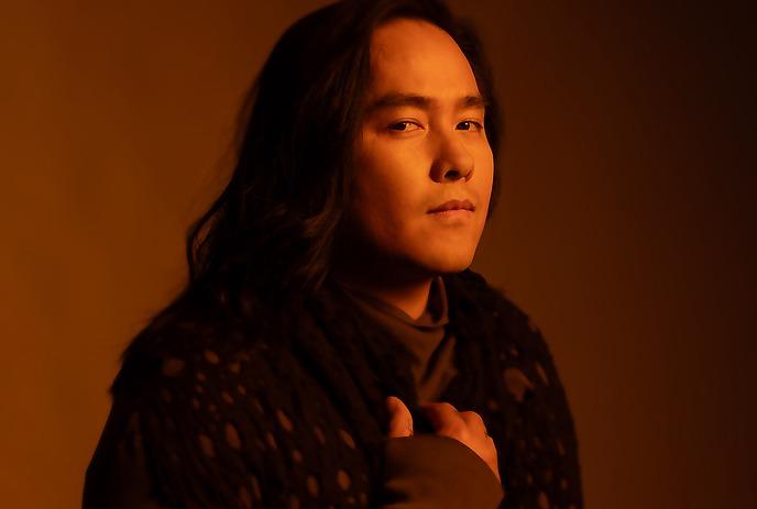 A person with a warm-toned background, wearing a dark, textured shawl and a long-sleeved shirt posing for the picture.