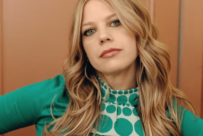 A close up of Basia Bulat, wearing a green dress against a brown background.