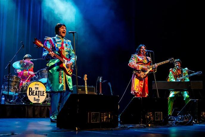 A lively band performs on stage, featuring a prominent large drum. The stage is illuminated with vibrant & colorful lights.