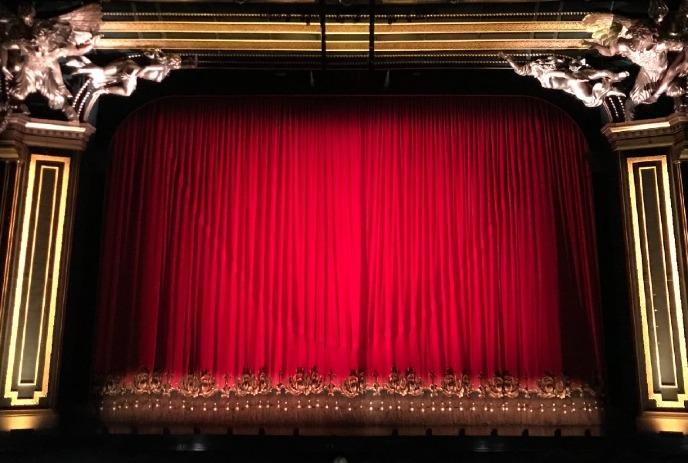 A theater stage with a closed red curtain, ornate golden proscenium arch, and intricate detailing.