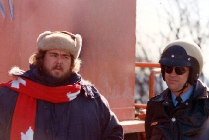 A man in a fur hat and Canada scarf stands beside a police officer with a helmet with pinkish wall in the background.