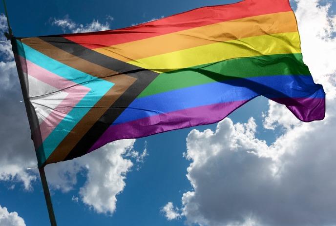 The pride flag waving in the wind on a sunny day, with clouds behind it.