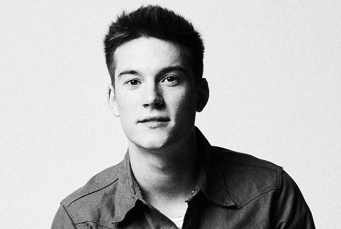 A black and white portrait of Cameron Whitcomb wearing a shirt with a gentle smile under a white background.