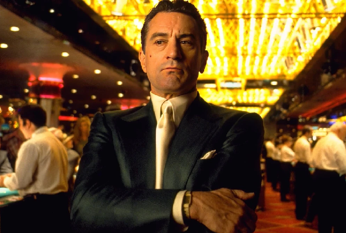 A man in a formal suit stands confidently in a lively casino with bright lights, engaged in activities at tables.