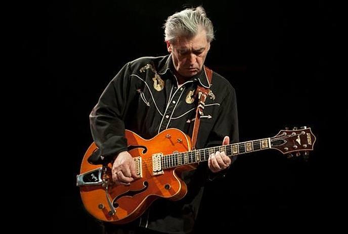 Chris Spedding playing his guitar, in front of a pitch black background.