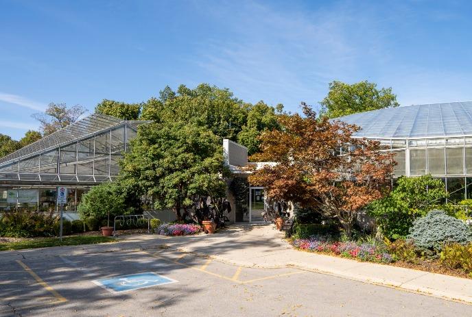 The image shows the exterior of a greenhouse building surrounded by vibrant greenery, trees, and colorful flowers.