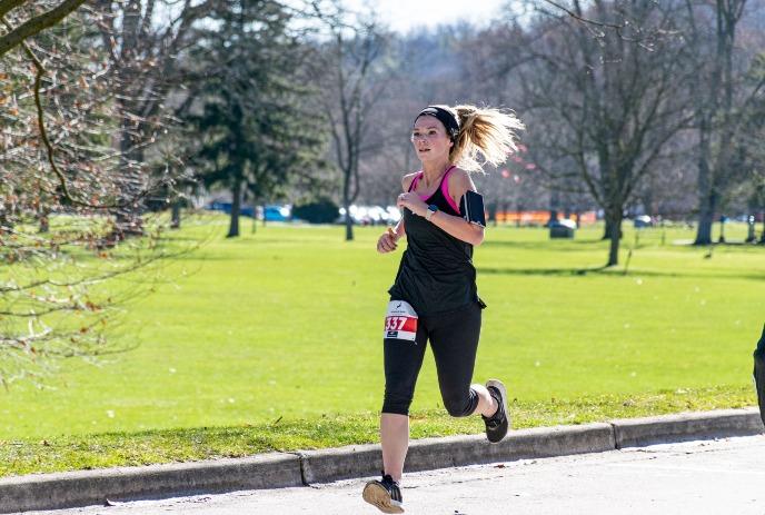 A woman running outdoors in a park, wearing black athletic clothing and a race bib numbered '437,' around trees.