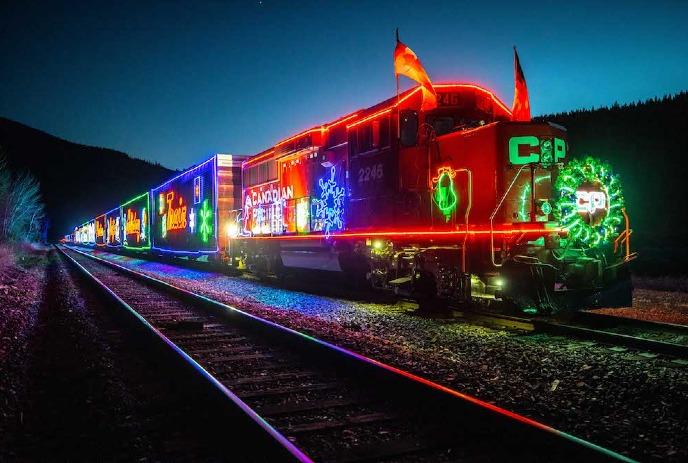 A brightly lit CPKC Holiday Train decorated with colorful lights and holiday designs travels along the tracks at night.