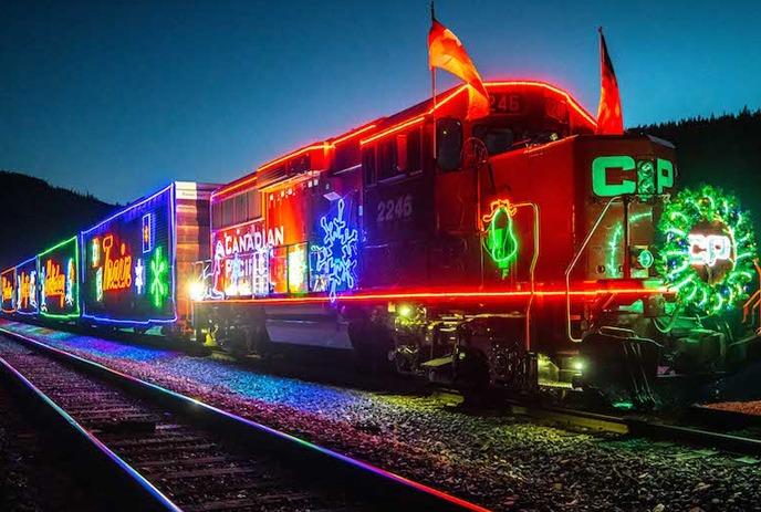 A brightly lit CPKC Holiday Train decorated with colorful lights and holiday designs travels along the tracks at night.