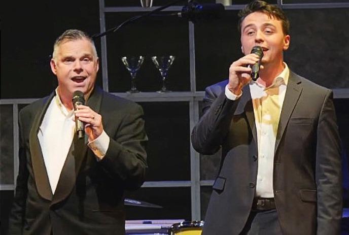 Two men in suits are singing on stage, positioned in the center of a background featuring black and white piano keys.