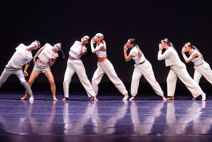 Seven dancers in white outfits perform a synchronized routine onstage under dramatic spotlight illumination.