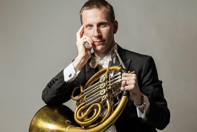 A person holding a French horn, wearing a white shirt and a wristwatch, against a black background.