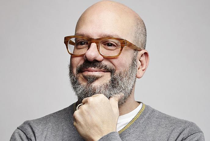 A portrait picture of David Cross wearing a black t-shirt and grey beard under a light yellow background