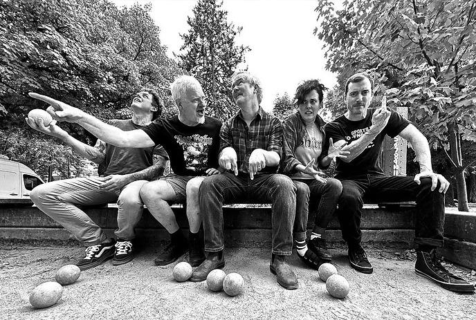 A grayscale photo of five people sitting on a bench in a park, surrounded by trees and greenery.