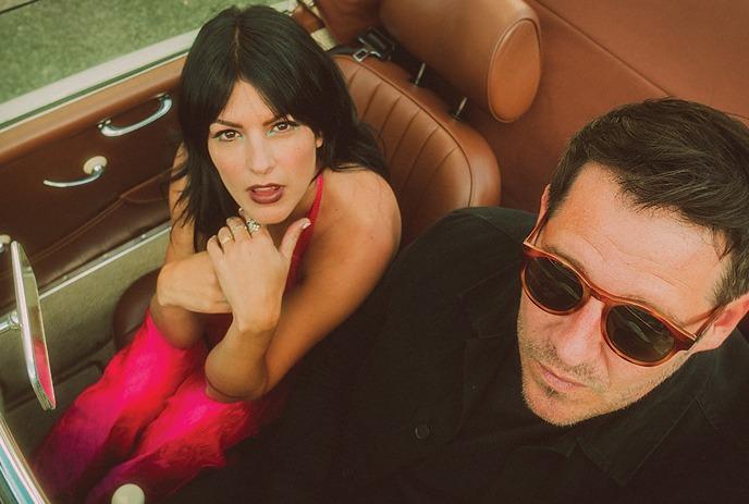 A man in black suite and glasses while a girl in a pink dress sitting in a convertible car and posing for a picture.