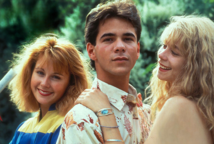 A young man with dark hair, wearing a patterned button-up shirt, standing between two smiling young women.