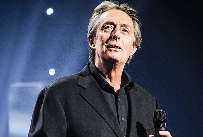 A comedian in a dark suit holds a microphone, gesturing mid-performance under dramatic blue stage lighting.