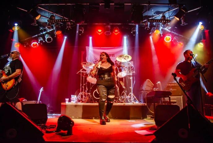 A live band performing on a stage with colored lights. Three members visible: singer with microphone, drummer, bassist.