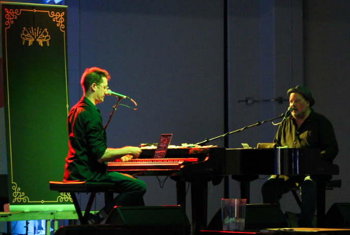 Two musicians are performing on stage with pianos under green spotlights.