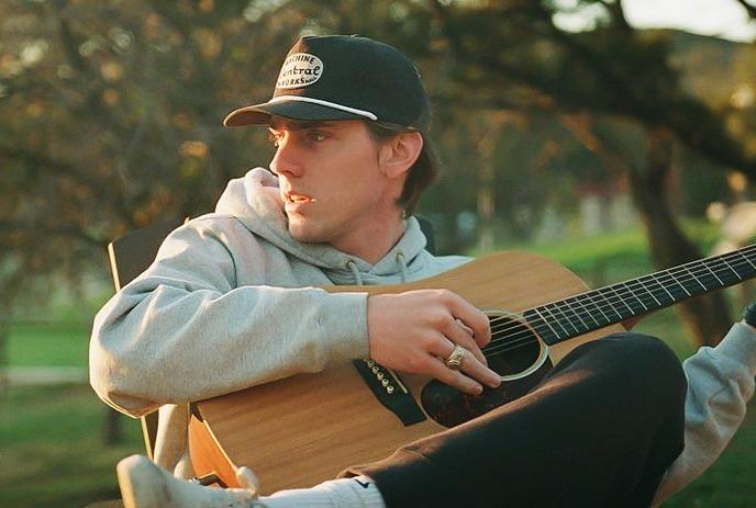 Dylan Gossett playing guitar outside in nature wearing a grey hoddie and a black hat.