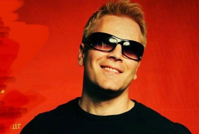 An artist in a black t shirt and black glasses smiling for the picture under a red background.