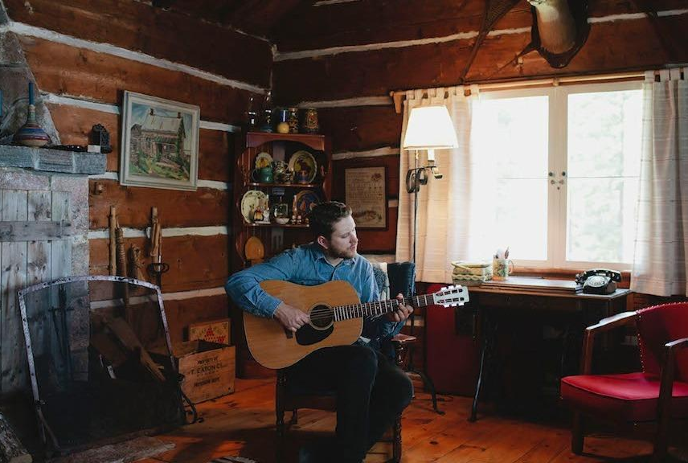 A person sitting in a cozy room playing an acoustic guitar, surrounded by wooden decor, a fireplace, a bookshelf.