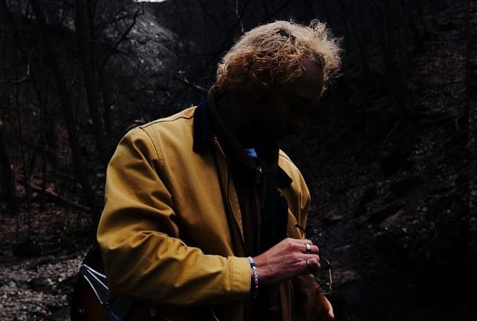 A person wearing a yellow jacket stands in a dimly lit forest or wooded area with a guitar.