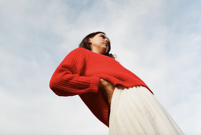 A woman in red sweater and white skirt standing against a clear sky looking straight, posing for the picture.