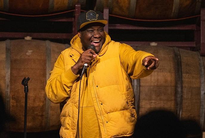 Gee Gethiga performing stand up comedy on stage, wearing a yellow jacket and a