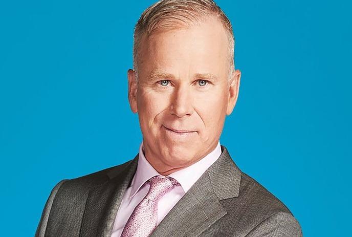 Gerry Dee posing in a suit in front of a blue background.