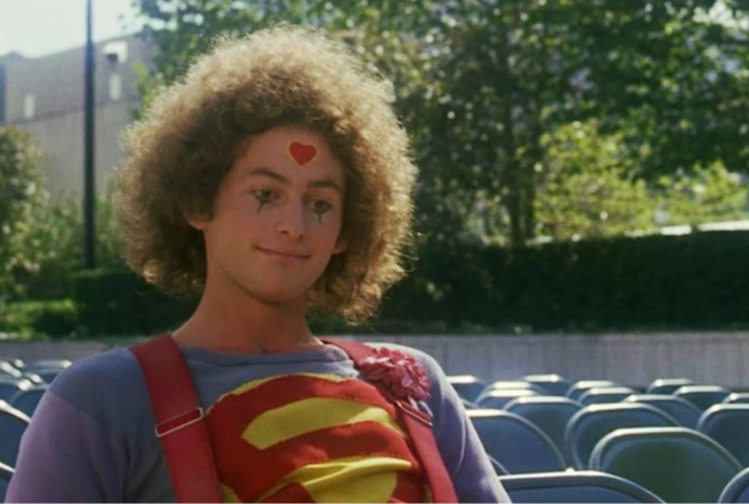 Victor Garber in a superman shirt and overalls, with poofy curly hair.