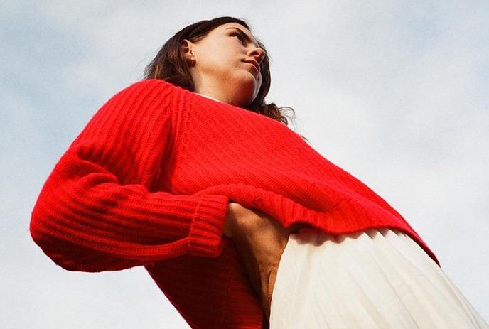 A woman in red sweater and white skirt standing against a clear sky looking straight, posing for the picture.