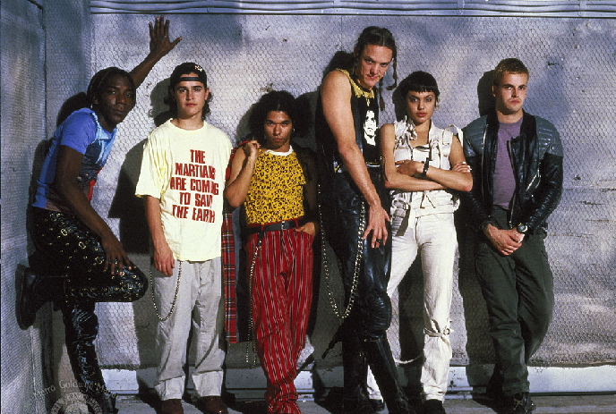 A group of six individuals posing against a wall with a chain-link fence, wearing diverse styles.