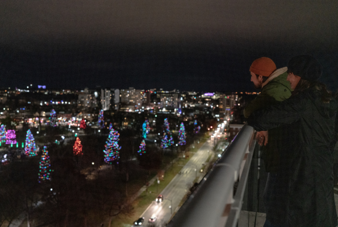 Two people stand on a balcony overlooking a cityscape at night, with streets and trees adorned with colorful holiday lights