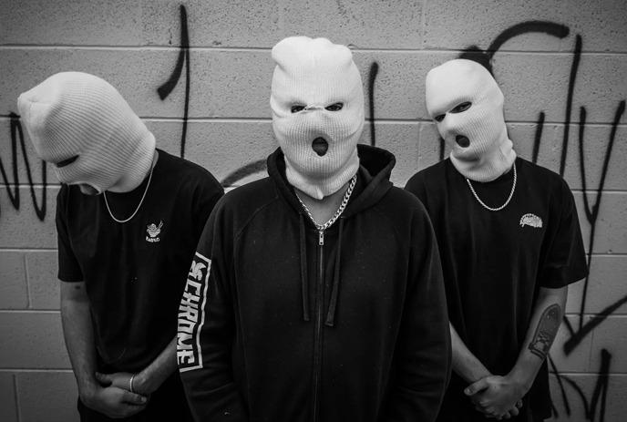 Three men standing in black outfits with white masks in a black and rusty background.