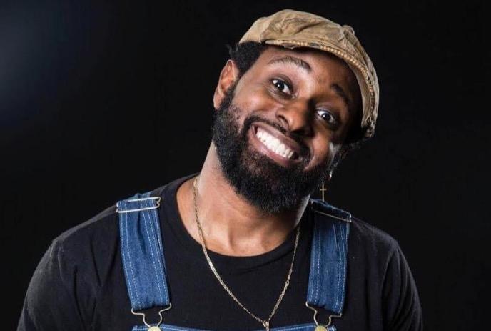 Jared Waters smiling and wearing a hat and a pair of overalls, against a black background.