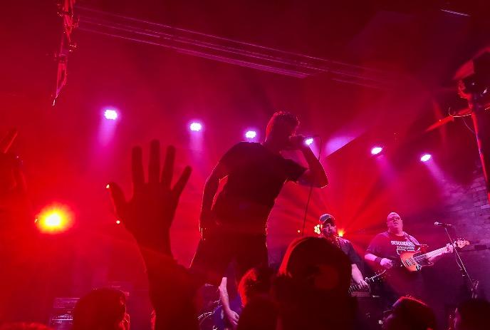 A band performing live on stage, surrounded by red lighting, with an engaged audience cheering.