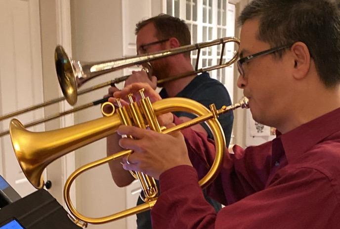 Two people playing brass instruments indoors; one plays a trombone, the other a flugelhorn.