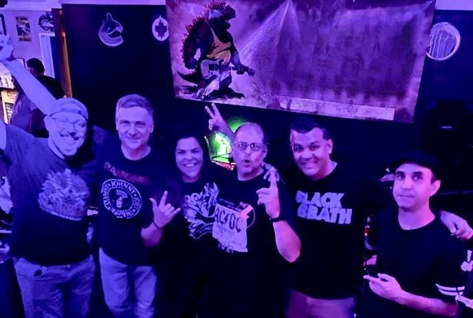 6 band members posing in black T shirts with dark background under purple and blue light and a poster with Brazilla.