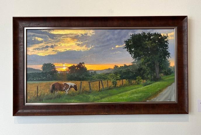 A framed painting of a horse grazing near a fence under a sunset sky.