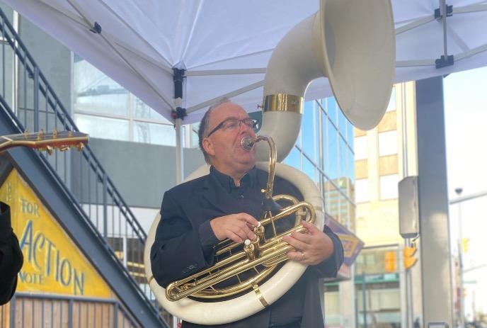 A person in a black suit is playing a sousaphone under a white canopy tent in an outdoor setting