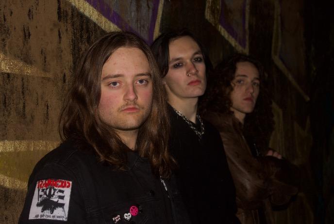 Three individuals stand against a wall, one wearing a black jacket with patches and pins posing for a picture.