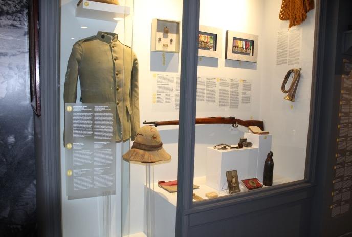 Museum exhibit with military artifacts: uniform jacket, helmet, rifle, medals, photos, and informational plaques.