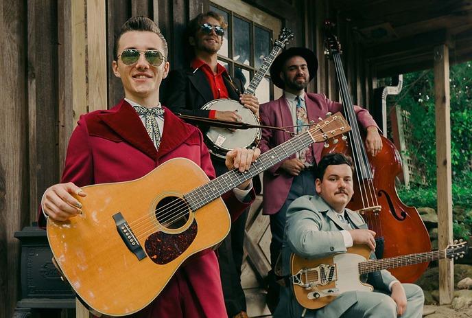 A group of four musicians poses outdoors with their instruments, including a guitar, banjo, bass, and electric guitar.