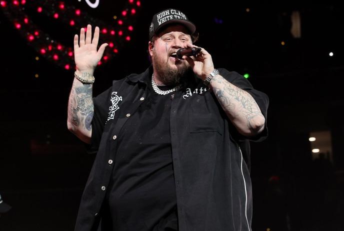 A tattooed performer on stage wearing a black button-up shirt and a hat with text, holding microphone and raising one hand.