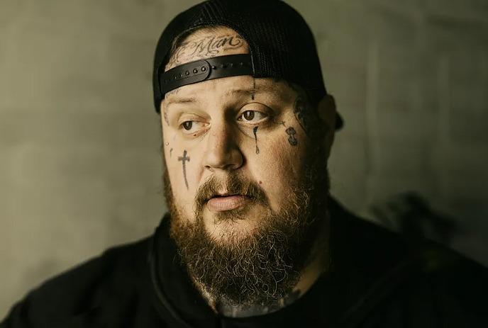 A close up of Jelly Roll, wearing a black hat and shirt, looking sideways to the camera.