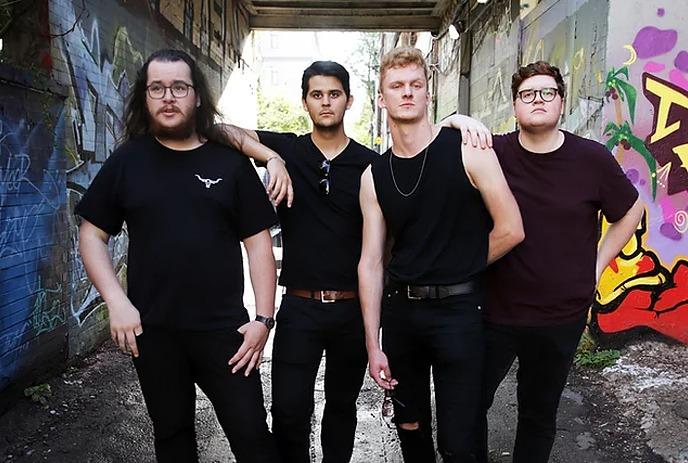 Four bandmembers standing in an alleyway with graffiti-covered walls, dressed in casual dark clothing posing for the picture