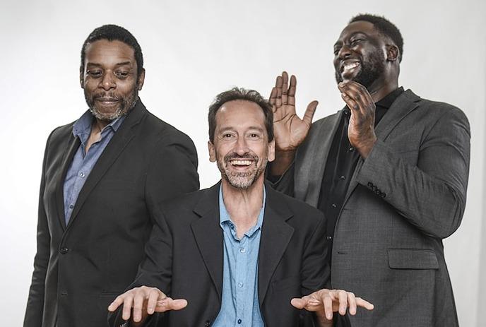 Three men in suits stand together, two of them smiling and one laughing with hands raised, set against a yellow background.