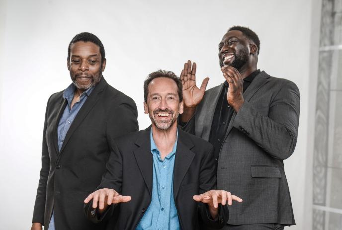 Three men in suits stand together, two of them smiling and one laughing with hands raised, set against a yellow background.